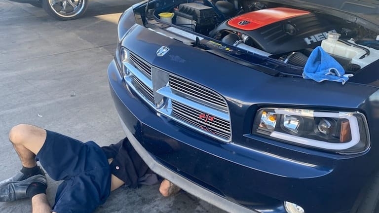 Mechanic lying under the front bumper of a navy Dodge Charger with the hood open, orange HEMI engine cover showing and a blue towel resting on the fender